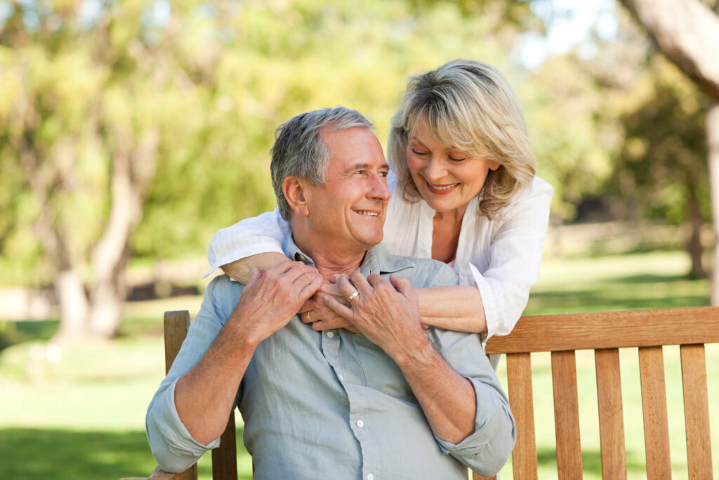 Senior,Woman,Hugging,Her,Husband,Who,Is,On,The,Bench